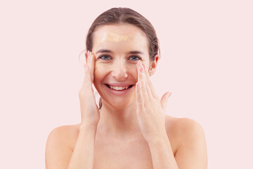 Naklejka premium Smiling young woman with natural skin applying foundation cream on face. Visible makeup strokes. Beauty portrait, isolated on pink background. Skincare and cosmetics routine, natural look concept.