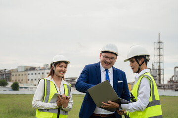 Executive Manager with Engineering Team Discussing Project on Laptop at Industrial Site. Three people are standing in a field, one of them is holding a laptop
