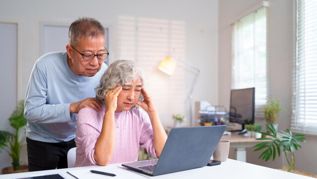 Stressed Asian Senior Woman with Headache Using Laptop, Husband Comforts. Support for Elderly Caregivers & Mental Health in Senior Couple at Home