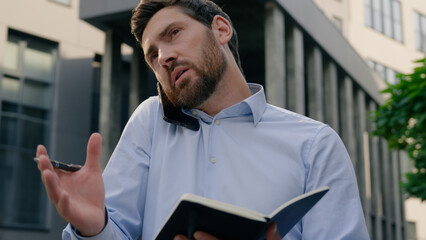 Serious male employer talking mobile phone call in city writing notes outdoors downtown write in...