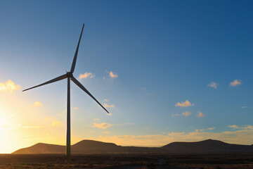 A large wind turbine stands against a colorful sunset with mountains in the background. The sky is...