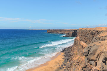 Dramatic cliffs overlook the turquoise waters and sandy beaches of Fuerteventura in the Canary Islands on a clear sunny day.