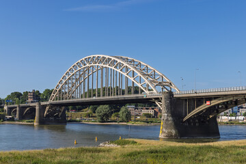 Obraz premium The grand Waalbrug, a prominent arch bridge, spans the Waal River in Nijmegen under a clear blue sky, connecting the vibrant city with its scenic surroundings on a bright day.
