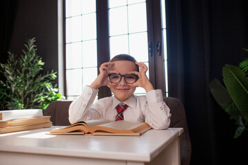 A 7-year-old child in glasses and  dressed school uniform poses at the school table indoor/ The boy learn the book