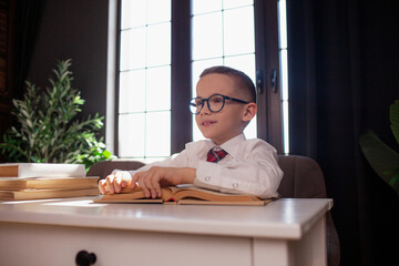 A 7-year-old child in glasses and  dressed school uniform poses at the school table indoor/ The boy learn the book