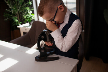 A 7-year-old child in glasses and dressed school uniform poses at the school table indoor. The boy looks to the microscope