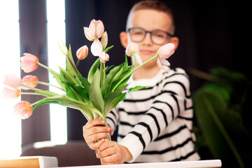 A 7-year-old child in glasses and dresses striped vest, poses at the school table indoor. The boy holds flowers
