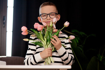 A 7-year-old child in glasses and dresses striped vest, poses at the school table indoor. The boy holds flowers