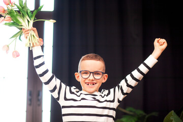 A 7-year-old child in glasses and dresses striped vest, poses at the school table indoor. The boy holds flowers