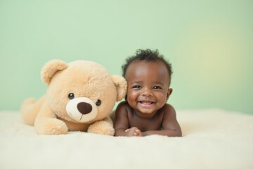 A Black newborn baby lies on a bed next to a large plush teddy bear, smiling with joy. The background is soft pastel mint green with warm light