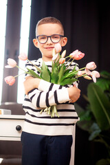A 7-year-old child in glasses and dresses striped vest, poses at the school table indoor. The boy holds flowers