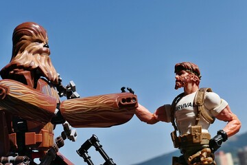 Naklejka premium LEGO Star Wars action figure of Wookie Chewbacca is going to shake hand (or paw) with GI JOE action figure of survival instructor Outback, Classified edition 2023. Blue skies background.