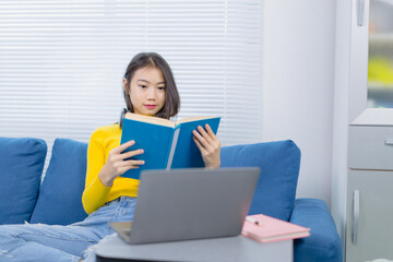 Asian woman reading a book and using laptop while sitting on comfortable blue sofa, studying online...