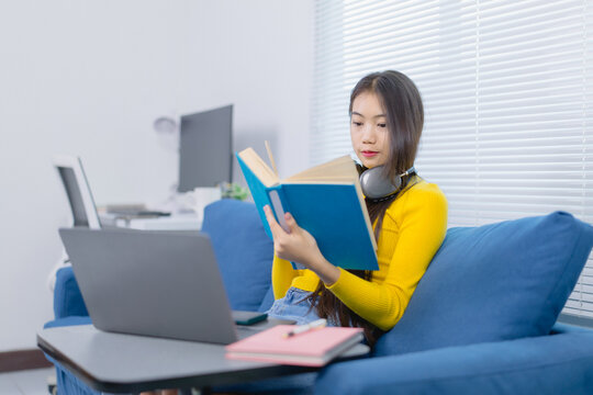 Asian female student is reading a book and using laptop for online learning at home during quarantine caused by the coronavirus pandemic, concept of online education and learning at home