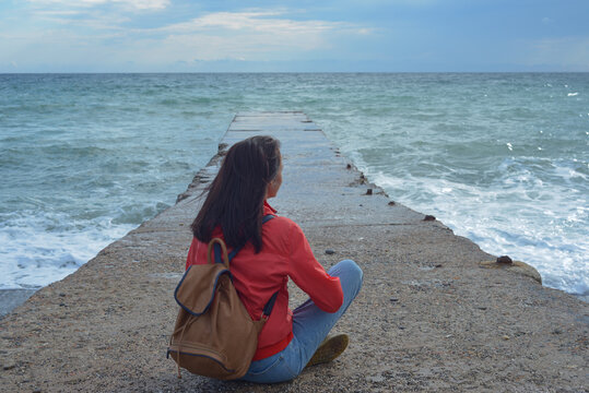 Rear view of a young woman tourist with a backpack sitting on a pier and waves hitting the pier in a storm. City of Sudak, Crimea.