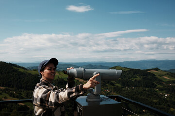 Woman points into the distance while looking through public binoculars on a scenic viewpoint in...