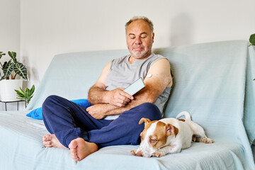 Mature man with diabetes and continuous glucose monitor on his arm using mobile app for checking his health, while sitting in sofa with Jack Russell Terrier dog at home