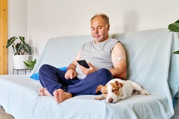 Mature man with diabetes and continuous glucose monitor on his arm using mobile app for checking his health, while sitting in sofa with Jack Russell Terrier dog at home