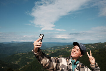 Happy woman shows peace sign while taking a selfie on a panoramic viewpoint in Serbian nature....