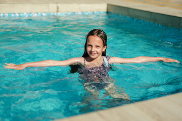 A happy young girl with outstretched arms enjoys a refreshing swim in a bright blue swimming pool...