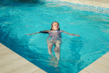 A young girl with closed eyes floats peacefully on her back in a bright blue swimming pool,...