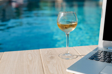 Glass of white wine and laptop on wooden poolside deck, symbolizing remote work, relaxation, and summer lifestyle by the water.