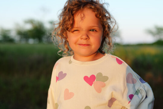 Smiling curly-haired girl in a heart-print sweatshirt stands in a sunny field, squinting in golden evening light with a playful expression.