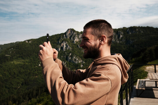 Side view of a young man smiling while taking a photo with his smartphone in the Serbian mountains. Male travel blogger