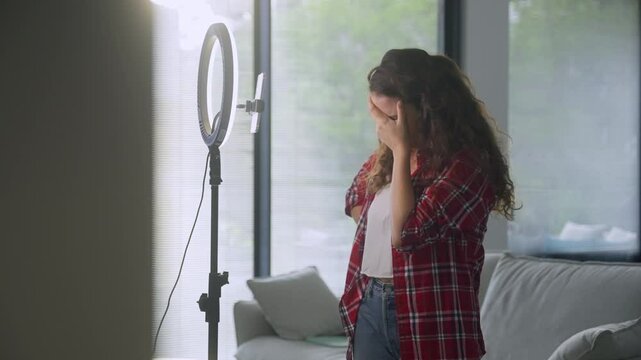 Nervous and angry woman preparing to go online for social content video creating. Female influencer making rant reel or personal vlog post speaking to phone on ring light stand in her home apartment