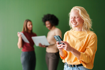 Happy Blonde Woman In Bright Yellow Sweater Texting On Phone While Friends Review Documents In...
