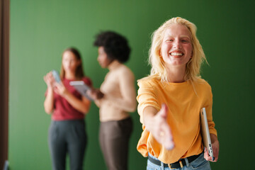 Smiling Blonde Woman Extending Hand Indoors with Diverse Group of Young Adults Using Technology in Background
