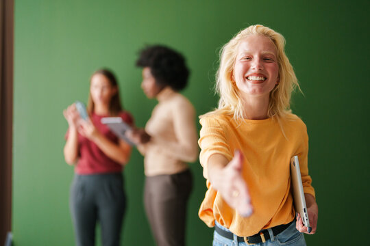 Smiling Blonde Woman Extending Hand Indoors with Diverse Group of Young Adults Using Technology in Background