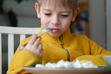 6 years old boy eats pasta with cheese from blue plate. Child is eating spaghetti. Lunch time. Good...