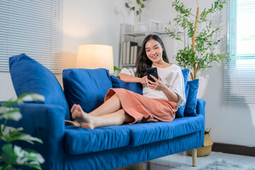 Obraz premium Happy asian woman using mobile phone while lying on comfortable sofa in living room at home