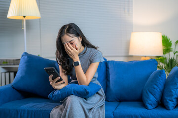 Stressed Asian woman is crying while using smartphone and sitting on sofa at home