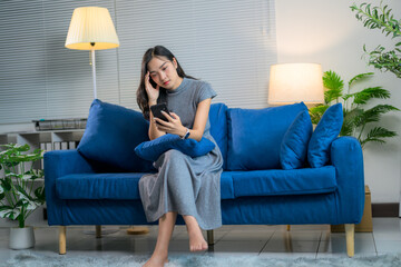 Young woman suffering from headache while using smartphone sitting on blue sofa in living room at...