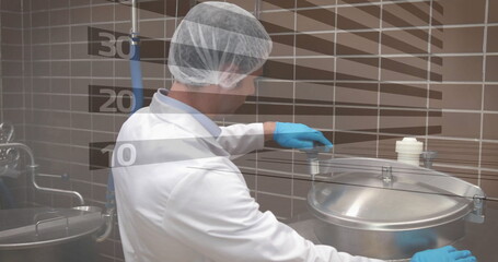 Technician in lab coat, gloves, hair net twisting latch on stainless steel tank in kitchen, pipes