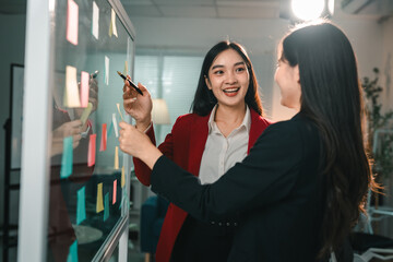 Two businesswomen brainstorming and discussing ideas using sticky notes on glass board in modern...