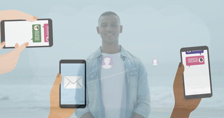 Standing man wearing denim shirt and white tee on sandy shore, with floating messaging smartphones