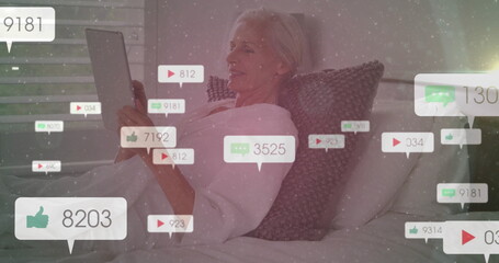 Reclining senior woman holding tablet computer in home bedroom, with floating social media icons