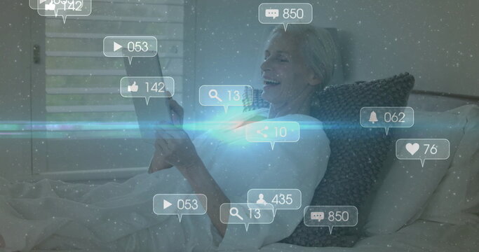 Reclining senior woman holding tablet on bed with gray woven pillow, with digital overlays floating - Powered by Adobe