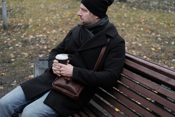 Pensive hipster drinking coffee in the urban street. Handsome man wearing black coat, brown leather...