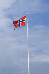 Photo with a view of the flag of Norway on a flagpole, fluttering in the wind, against a blue sky with clouds
