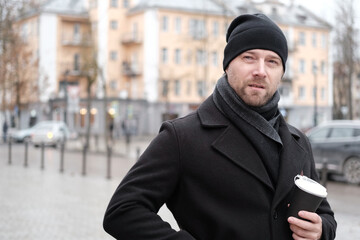 Pensive hipster drinking coffee in the urban street. Handsome man wearing black coat, brown leather...