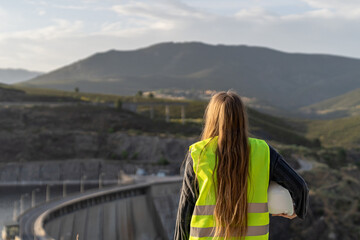 Female engineer standing with her back to the camera, holding her hardhat at her side,...