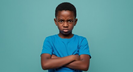 A young boy with a serious expression stands arms crossed against a teal backdrop expressing displeasure or defiance