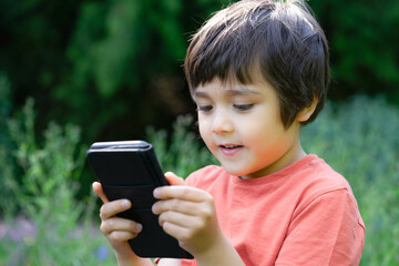 Happy kid boy sitting in the garden playing on a smartphone during playing outside in summer. Close up of adorable kid sitting alone holding a cell phone with blurry green forest nature