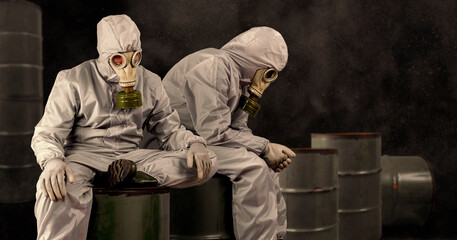 People in a gas masks and protective suits sitting on a barrel of chemical waste. Environmental pollution concept. Dirty air with dust and noise, grunge effect.