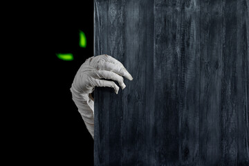 Mummy's hands wrapped in bandage rises through wooden boards. Background with copy space. Horrible glowing eyes stare out of the darkness of the risen dead mummy monster for Halloween horror concept.