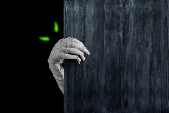 Mummy's hands wrapped in bandage rises through wooden boards. Background with copy space. Horrible glowing eyes stare out of the darkness of the risen dead mummy monster for Halloween horror concept. - Powered by Adobe